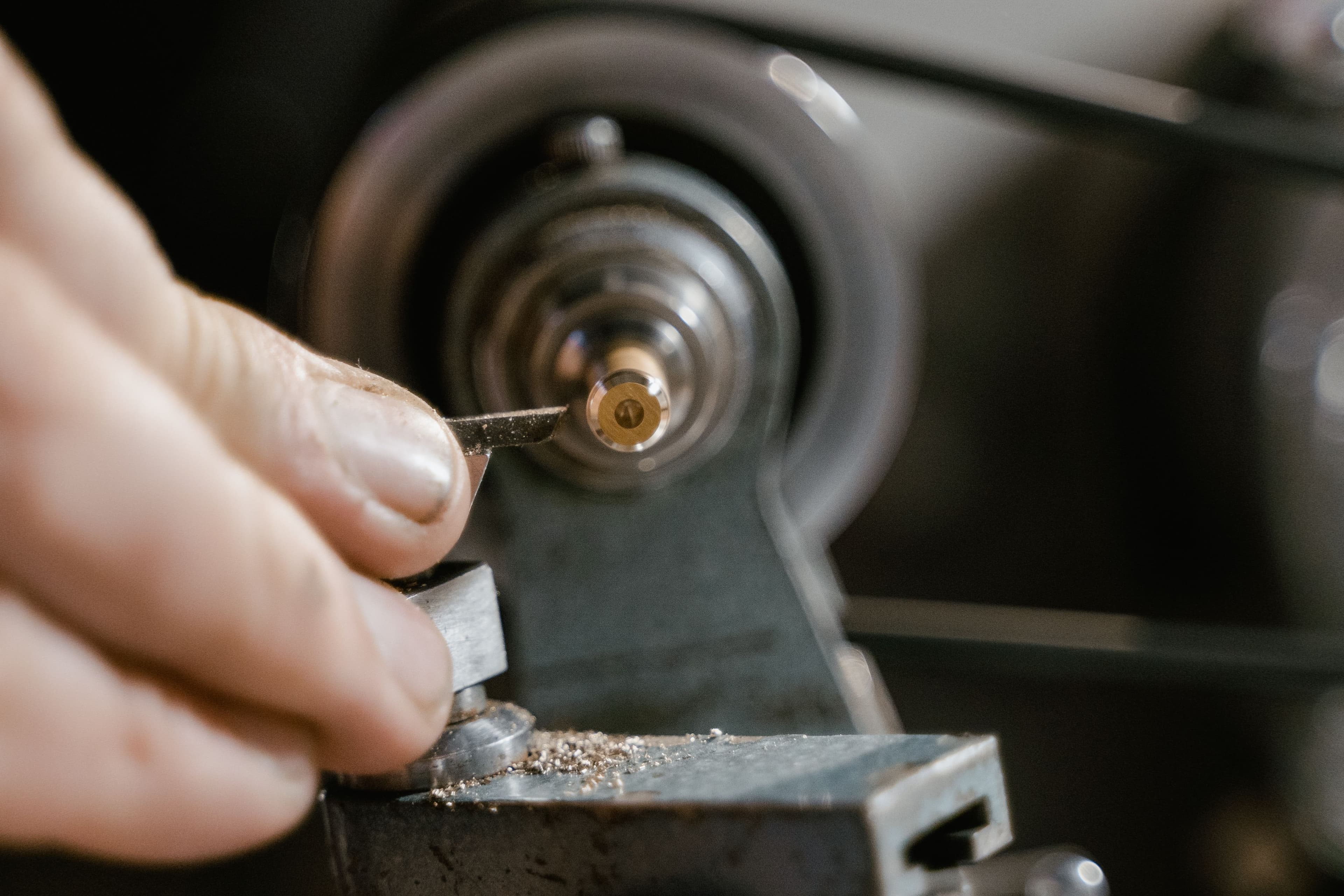 A gloved hand shapes a small brass part on a precision lathe.