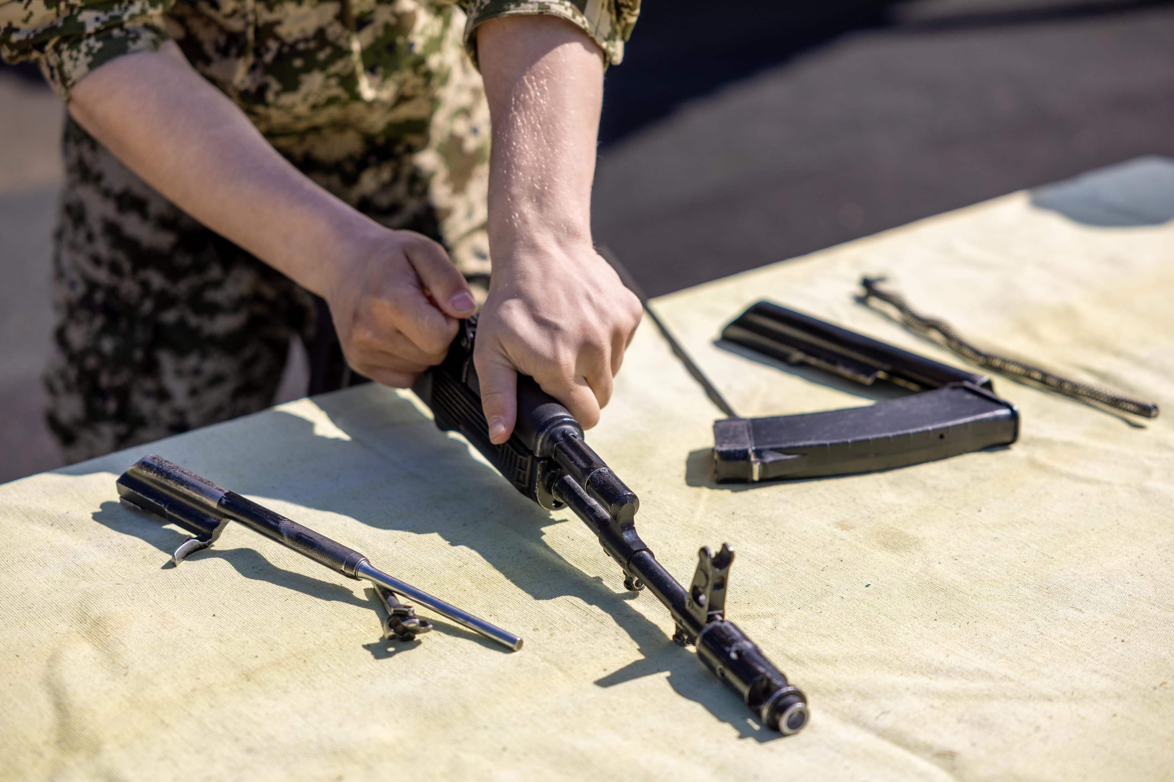 Hands disassembling a rifle into its component parts on an outdoor table.