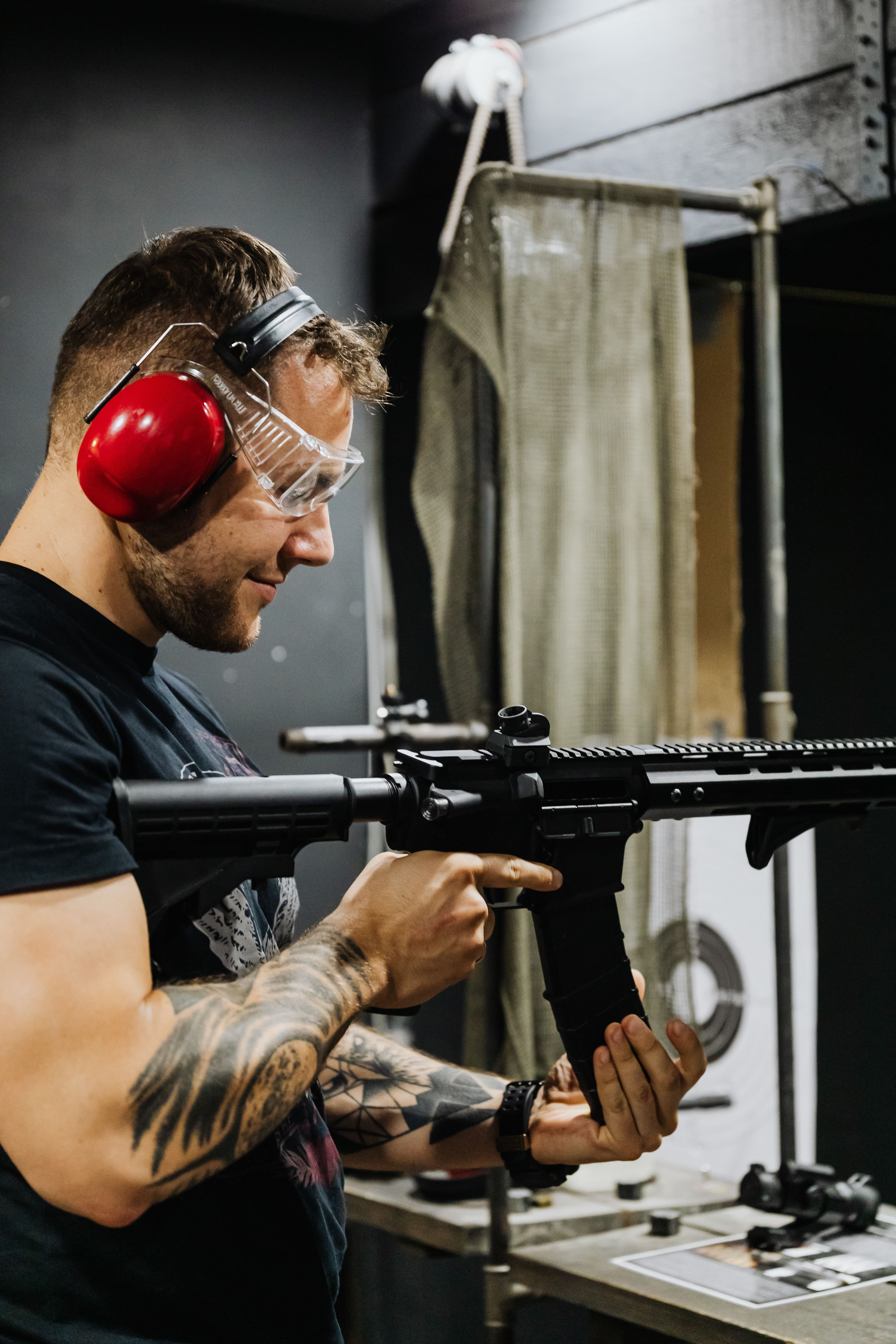 A shooter wearing ear and eye protection inspects a rifle at an indoor range.