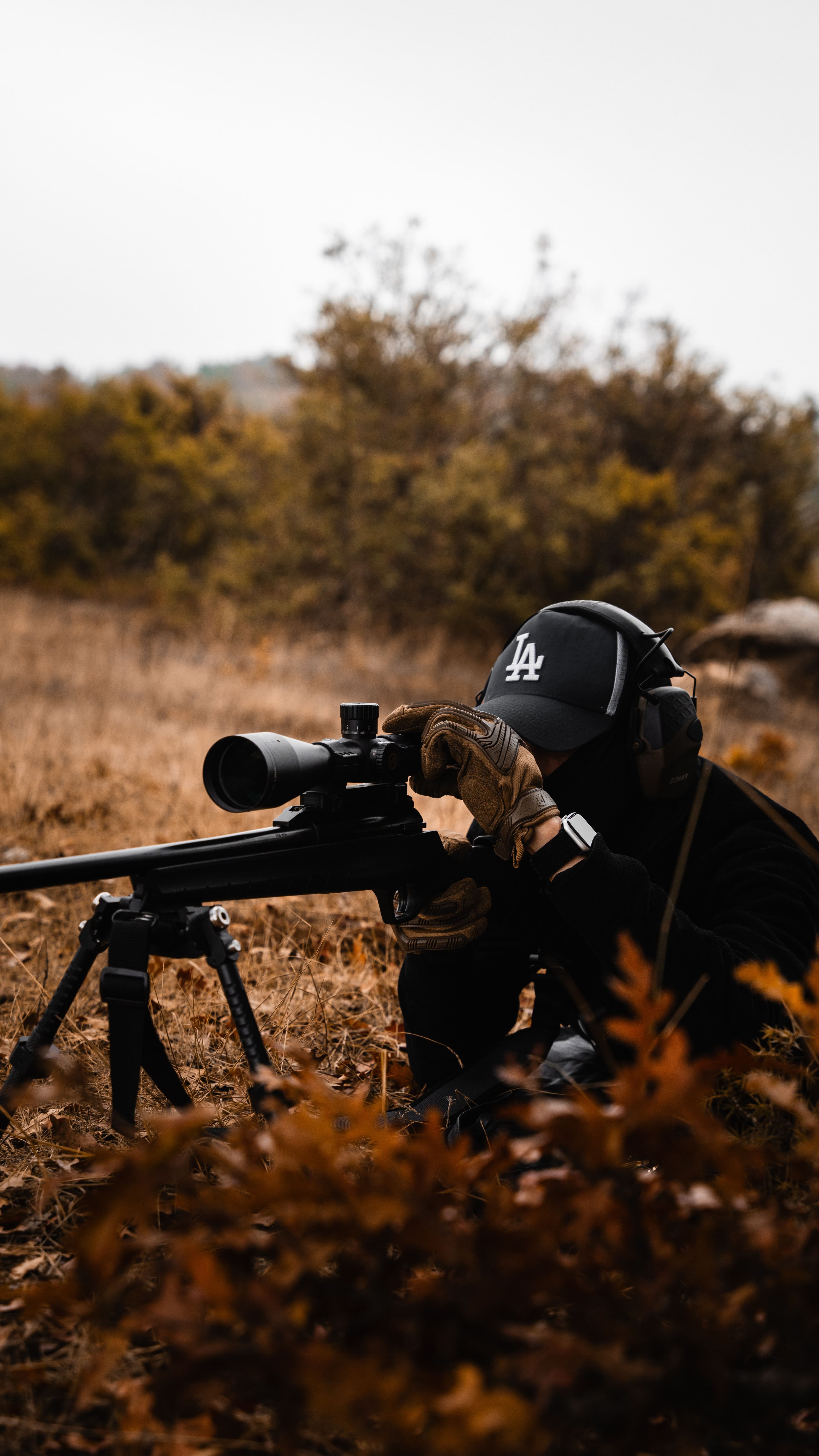 A shooter aims a bipod-mounted rifle through a scope in a field.