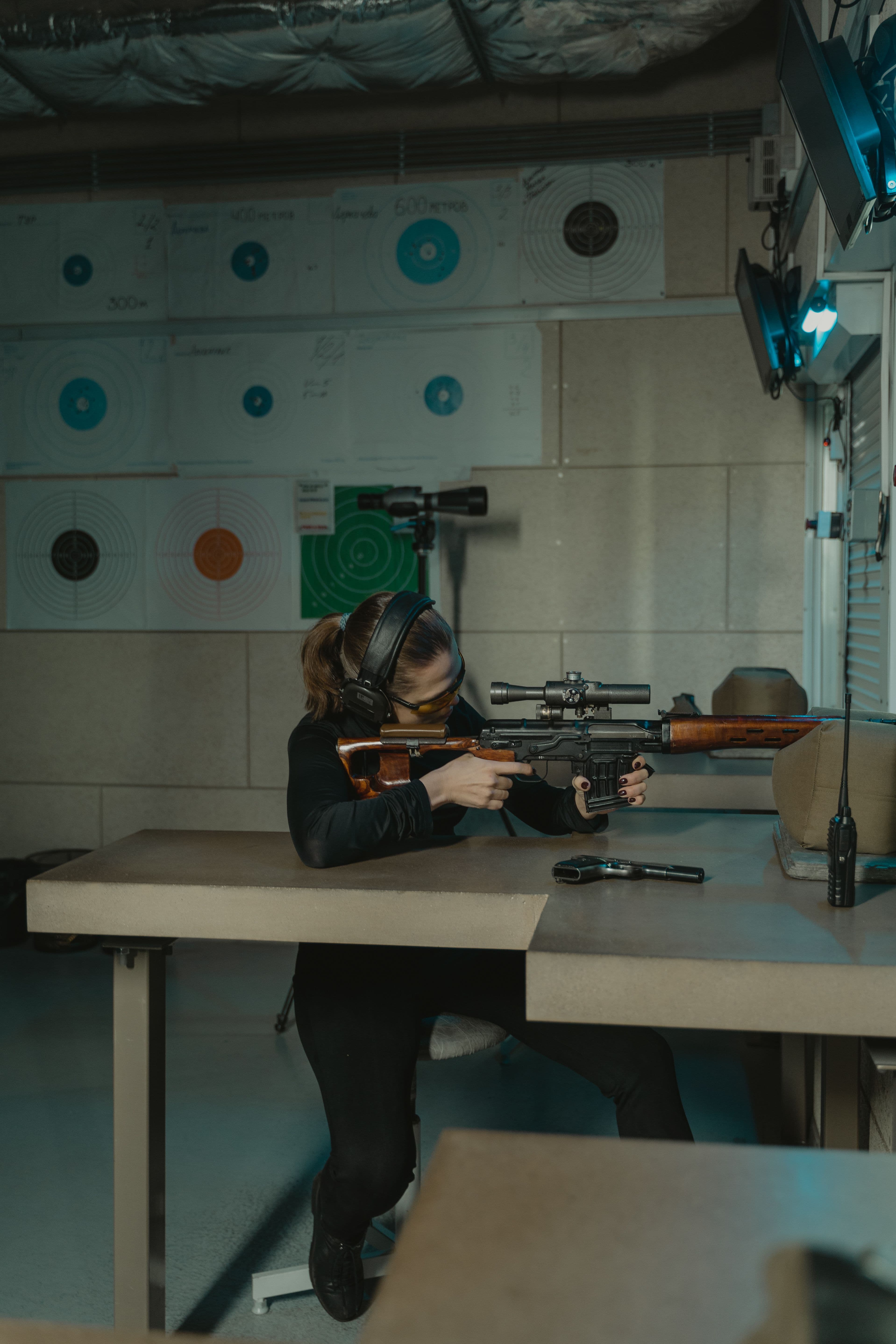 A shooter aims a wood-stocked, scope-mounted rifle at an indoor range.